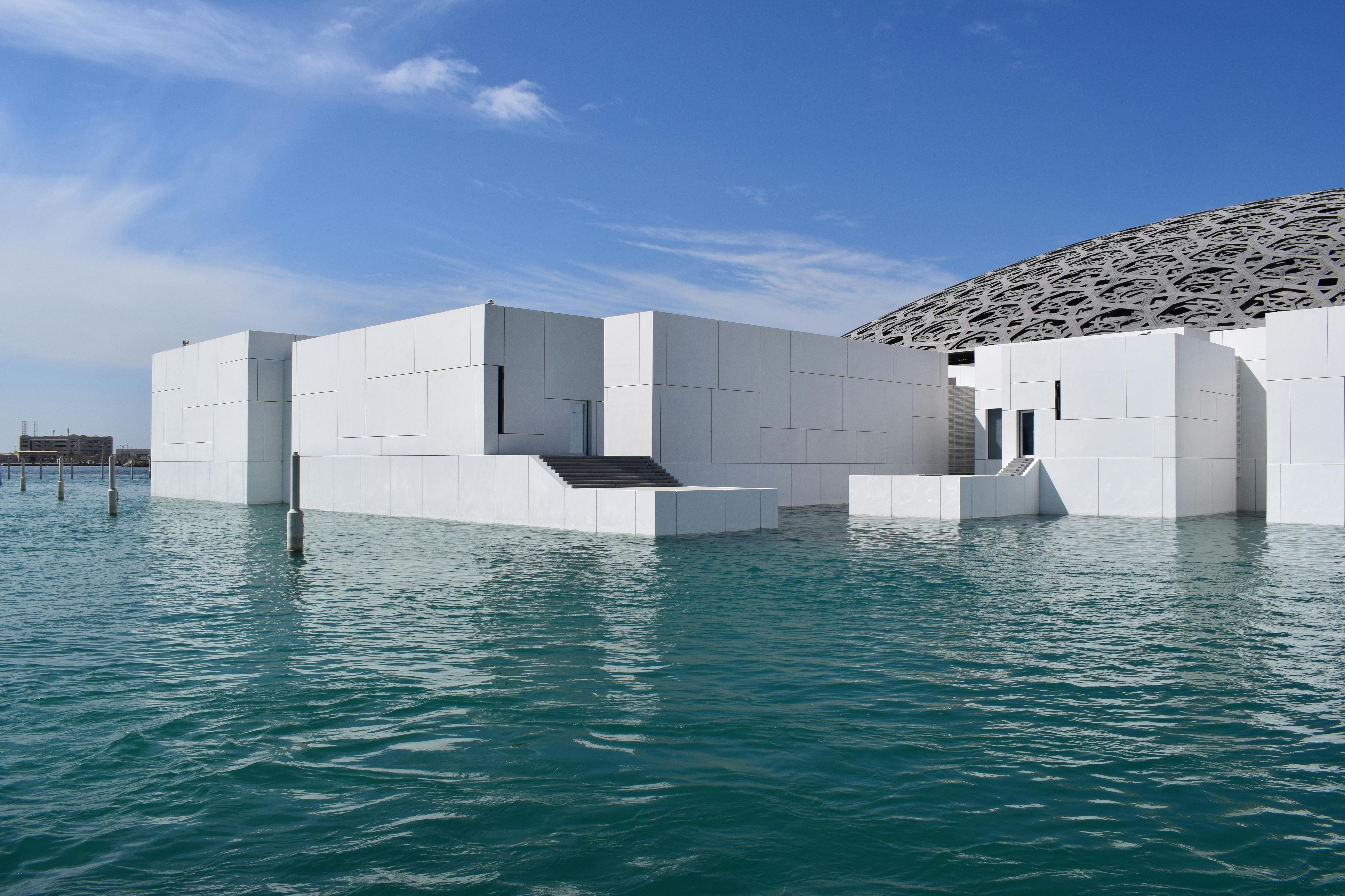 Modern white building with a geometric design and staircases, partially surrounded by water, under a clear blue sky.