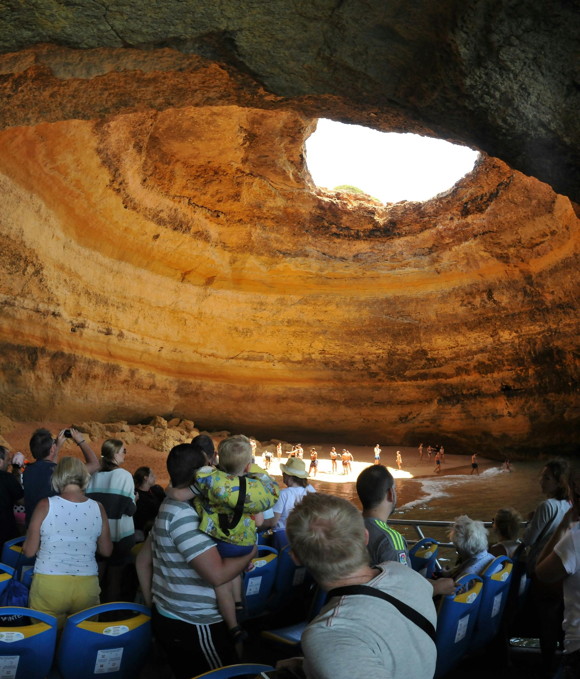 Tourists in a cave with a large circular opening at the top, viewing the sandy beach below. People take photos and enjoy the scene.