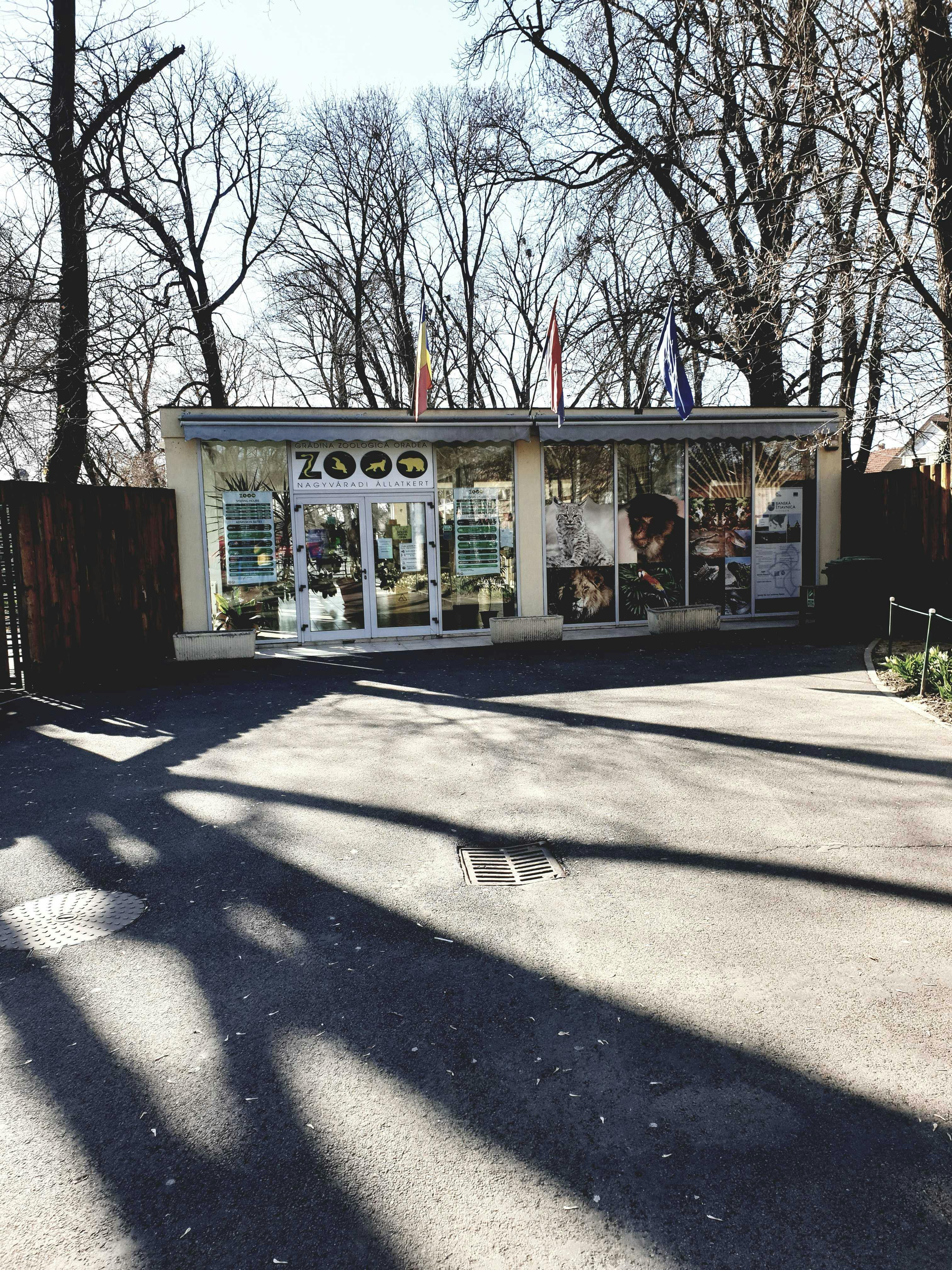 Single-story building with large windows, an entrance labeled "Zoo," and multiple flags on top. Leafless trees in the background.