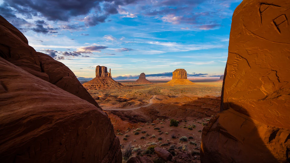 Three towering rock formations rise from an arid, desert landscape under a partly cloudy blue sky, viewed from a rocky vantage point.