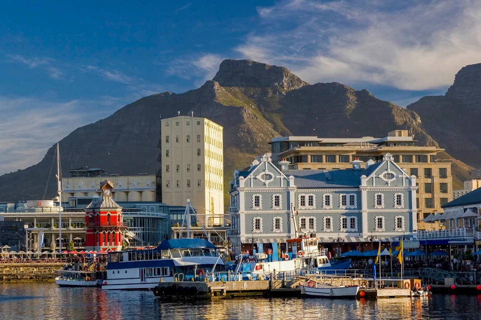 A waterfront with boats, colorful buildings, and a mountain in the background under a partly cloudy sky.