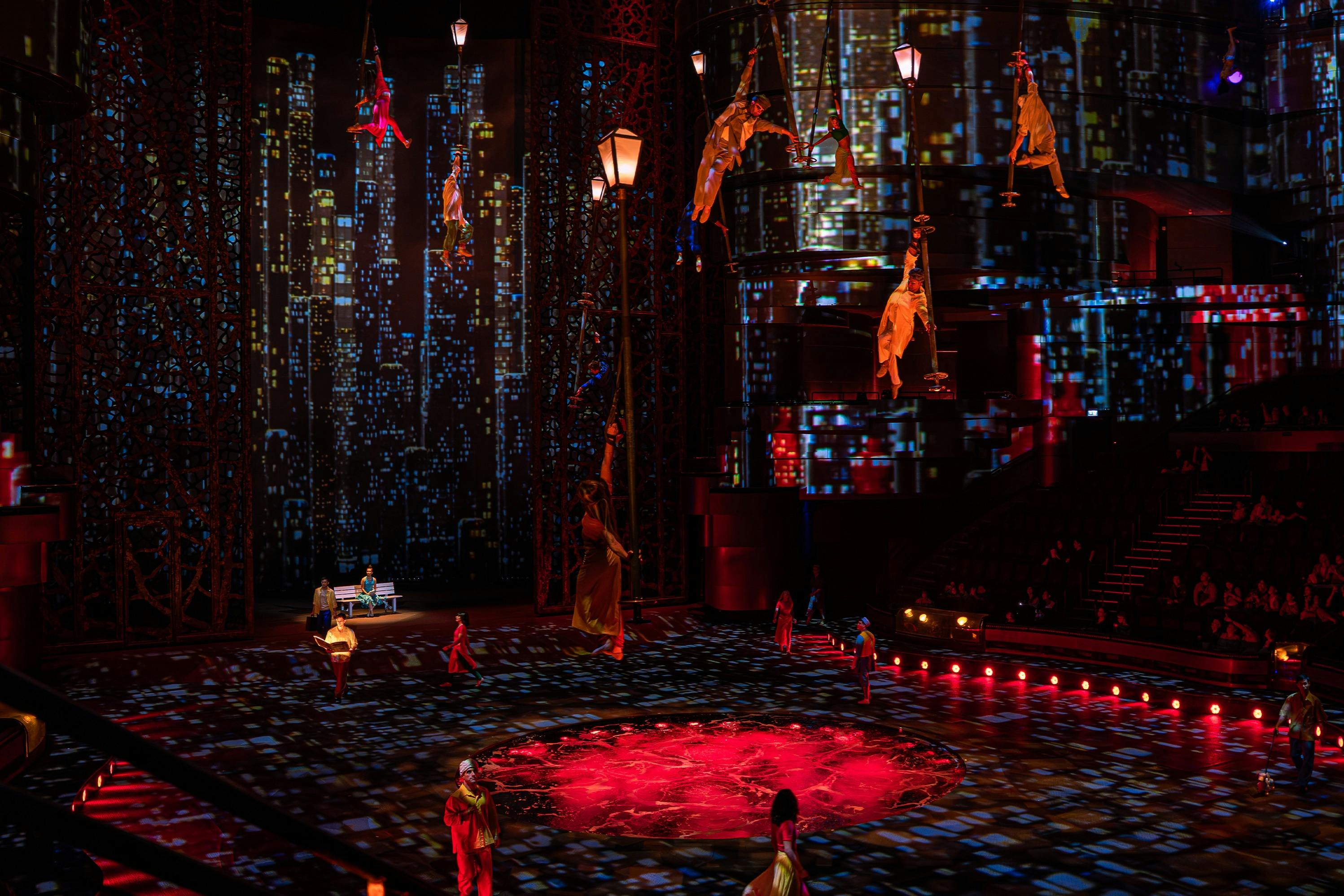 Performers in kleurrijke kostuums hangen aan touwen boven een verlicht podium met een achtergrond van een stadslandschap en zitplaatsen voor het publiek zichtbaar.