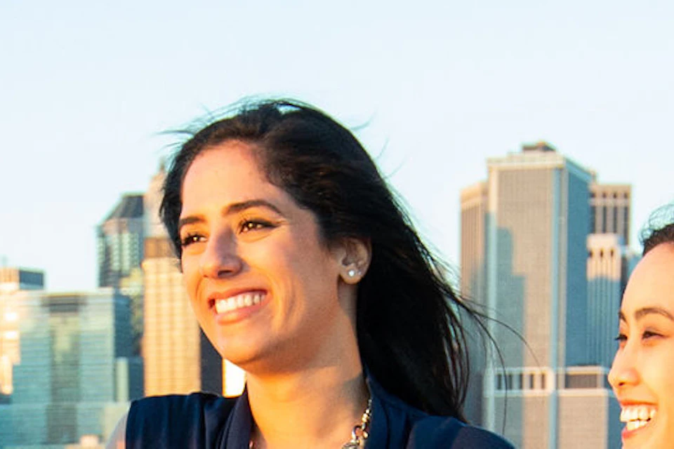 Four women stand on a waterfront pier, holding drinks and smiling. The city skyline is in the background under a clear sky.