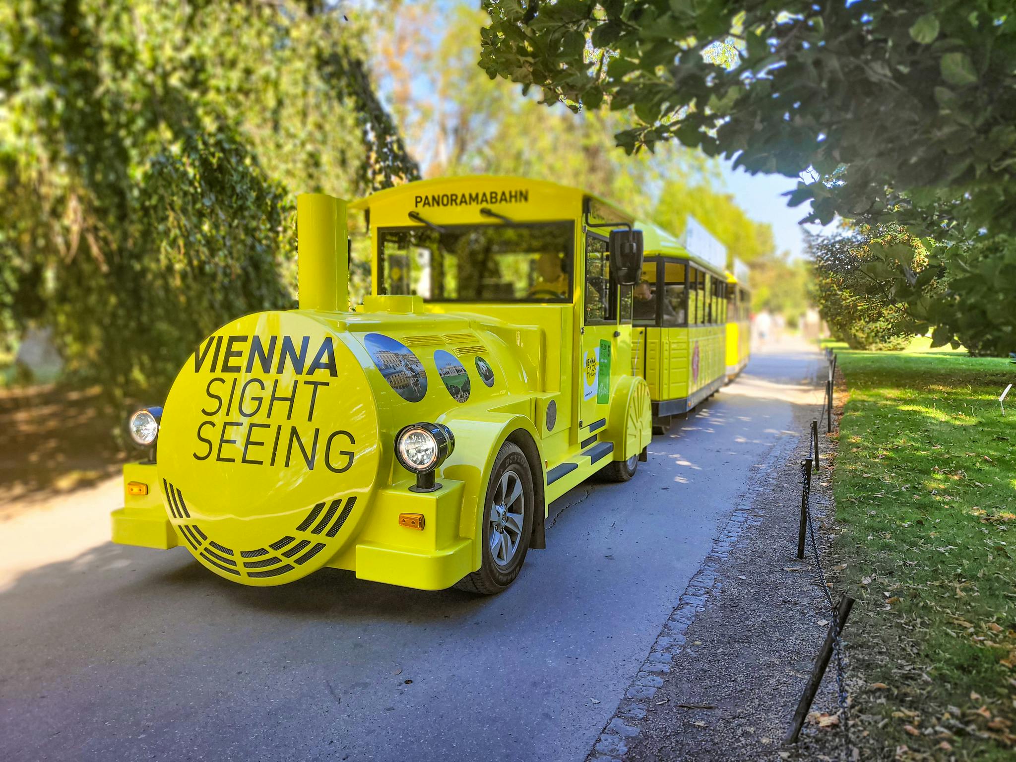 Ljusgul sightseeingspårvagn i en park märkt "Vienna Sightseeing" och "Panoramabahn", omgiven av frodig grönska.