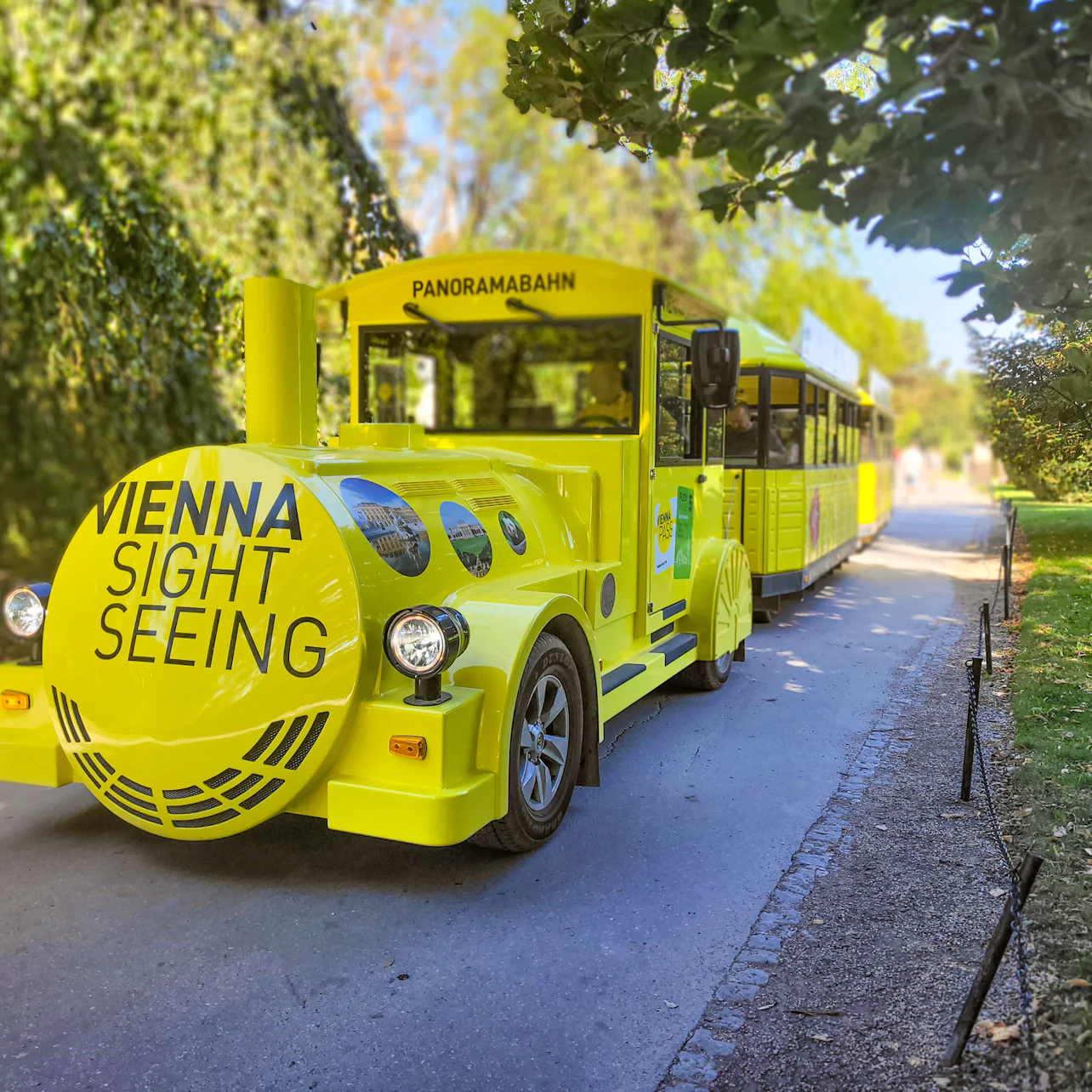 Bright yellow sightseeing tram in a park labeled "Vienna Sightseeing" and "Panoramabahn," surrounded by lush greenery.