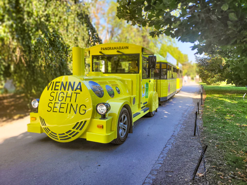 Bright yellow sightseeing tram in a park labeled "Vienna Sightseeing" and "Panoramabahn," surrounded by lush greenery.