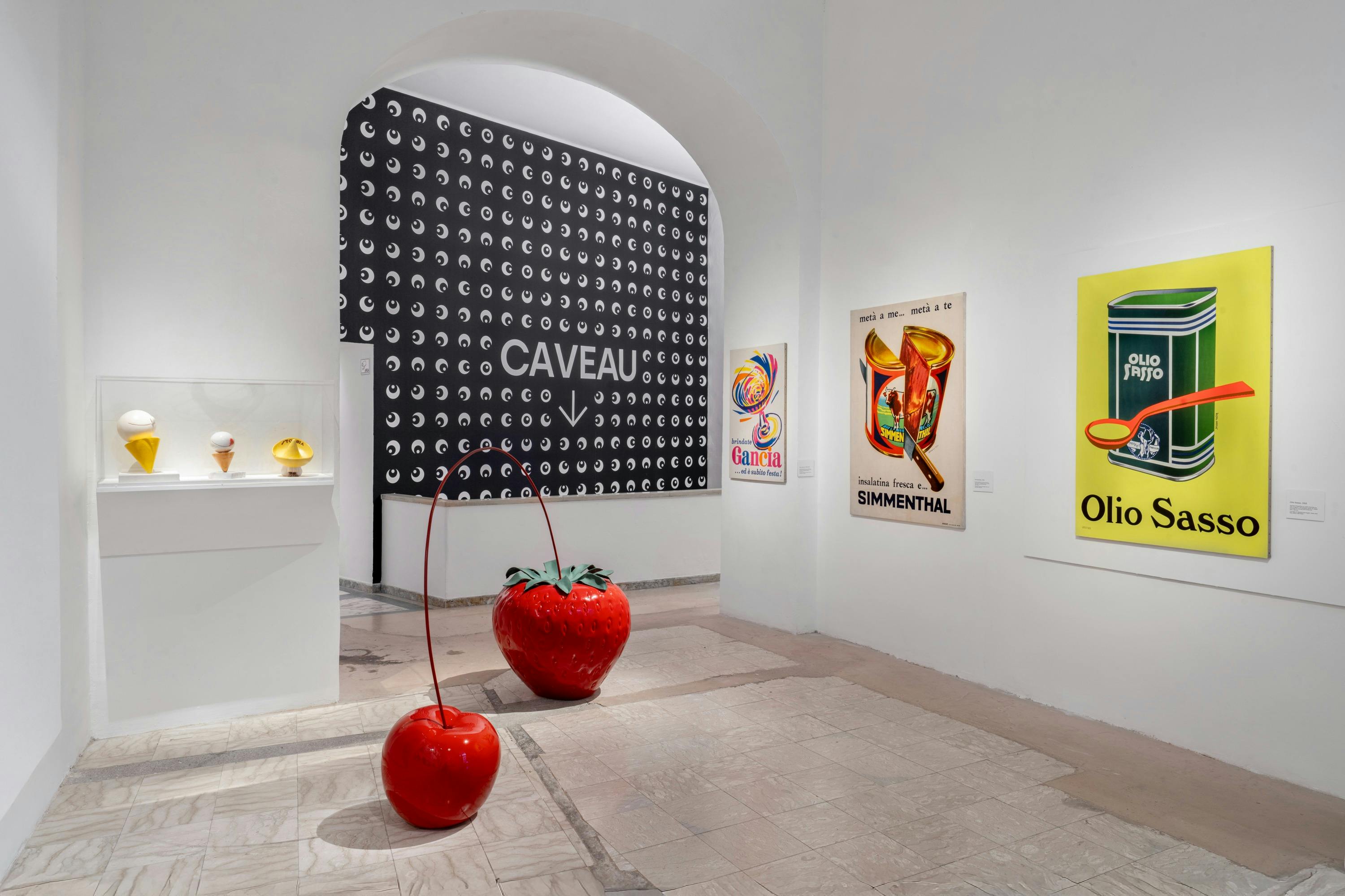 Ein Galerieraum mit Pop-Art-Postern und Skulpturen, darunter eine große rote Erdbeere und eine Kirsche auf dem Boden.
