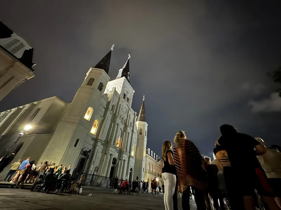 A nighttime view of a cathedral with illuminated windows and tall spires, with a group of people gathered in the foreground.