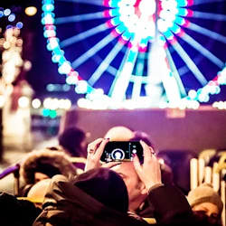 Person taking a photo with a smartphone, capturing a brightly lit Ferris wheel in the background, surrounded by a crowd at night.