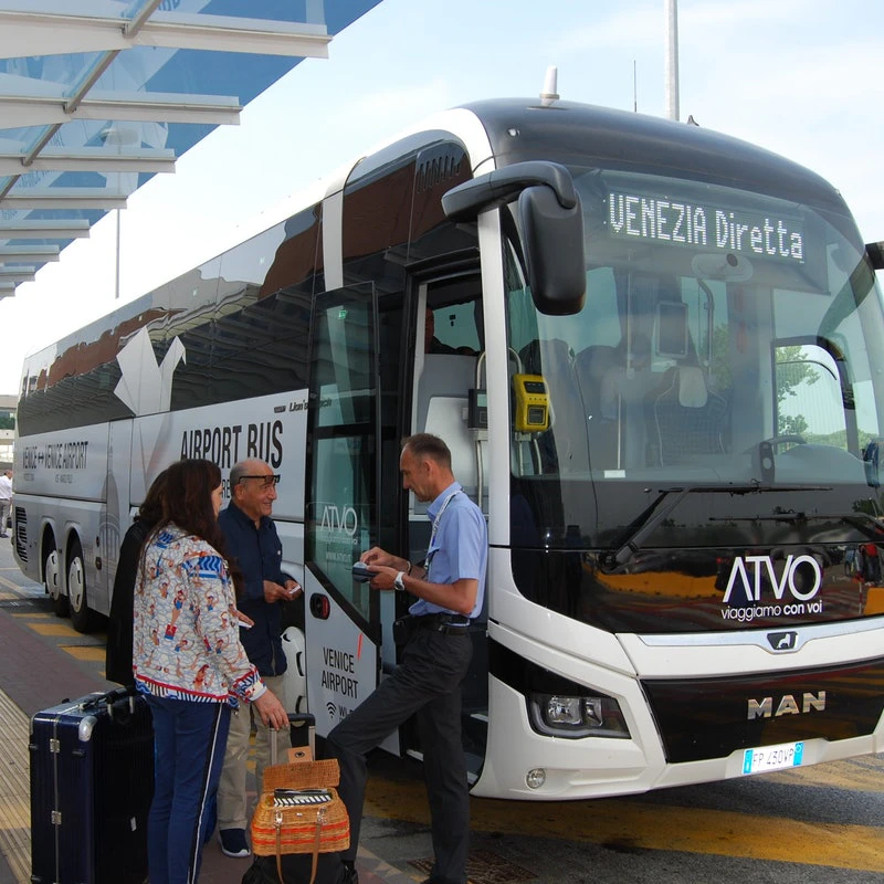 Three people standing outside a white "Venezia Diretta" airport bus, one of them boarding, with luggage nearby.