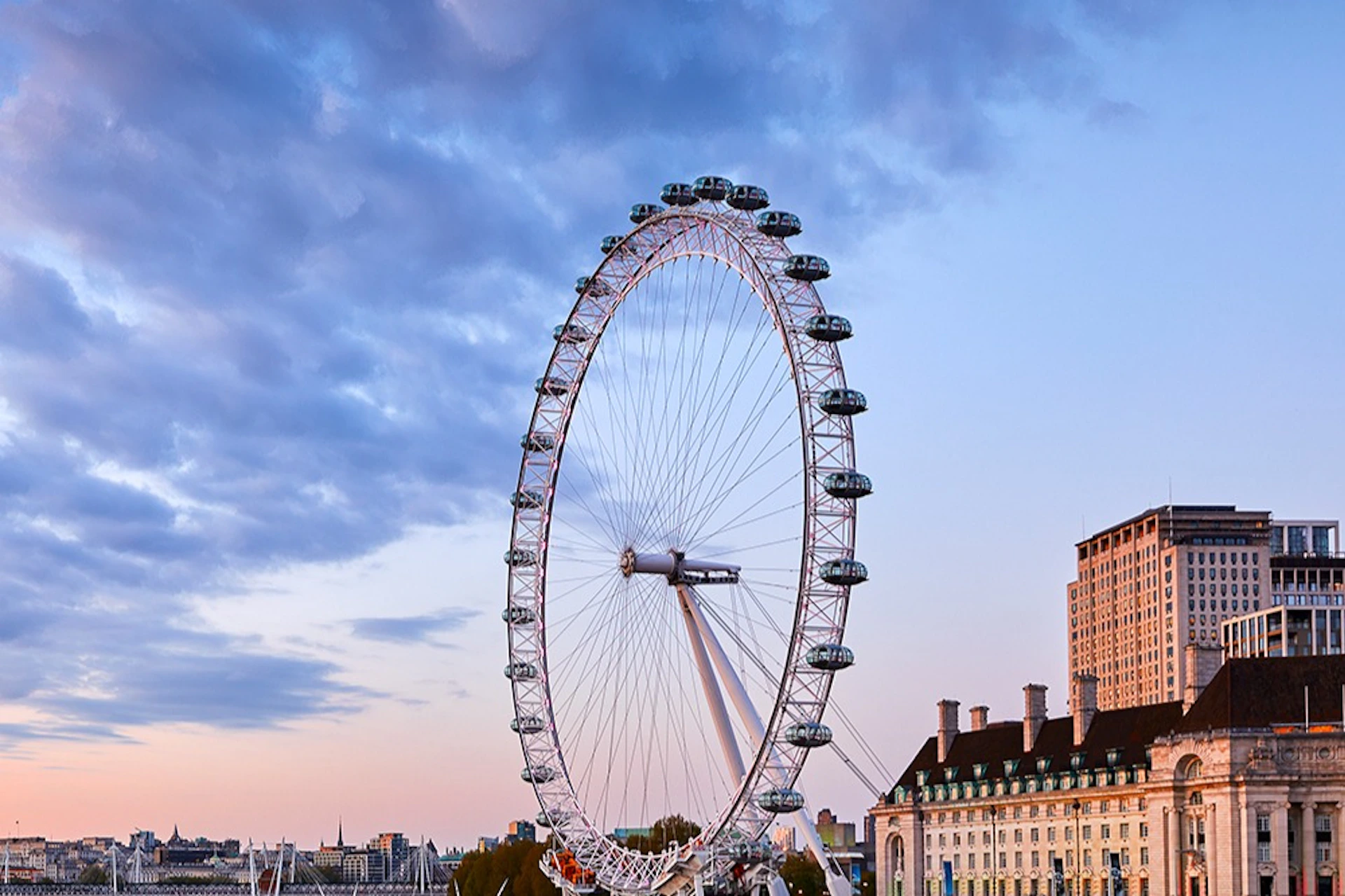 A large Ferris wheel, known as the London Eye, against a partly cloudy sky with buildings in the background at sunset.