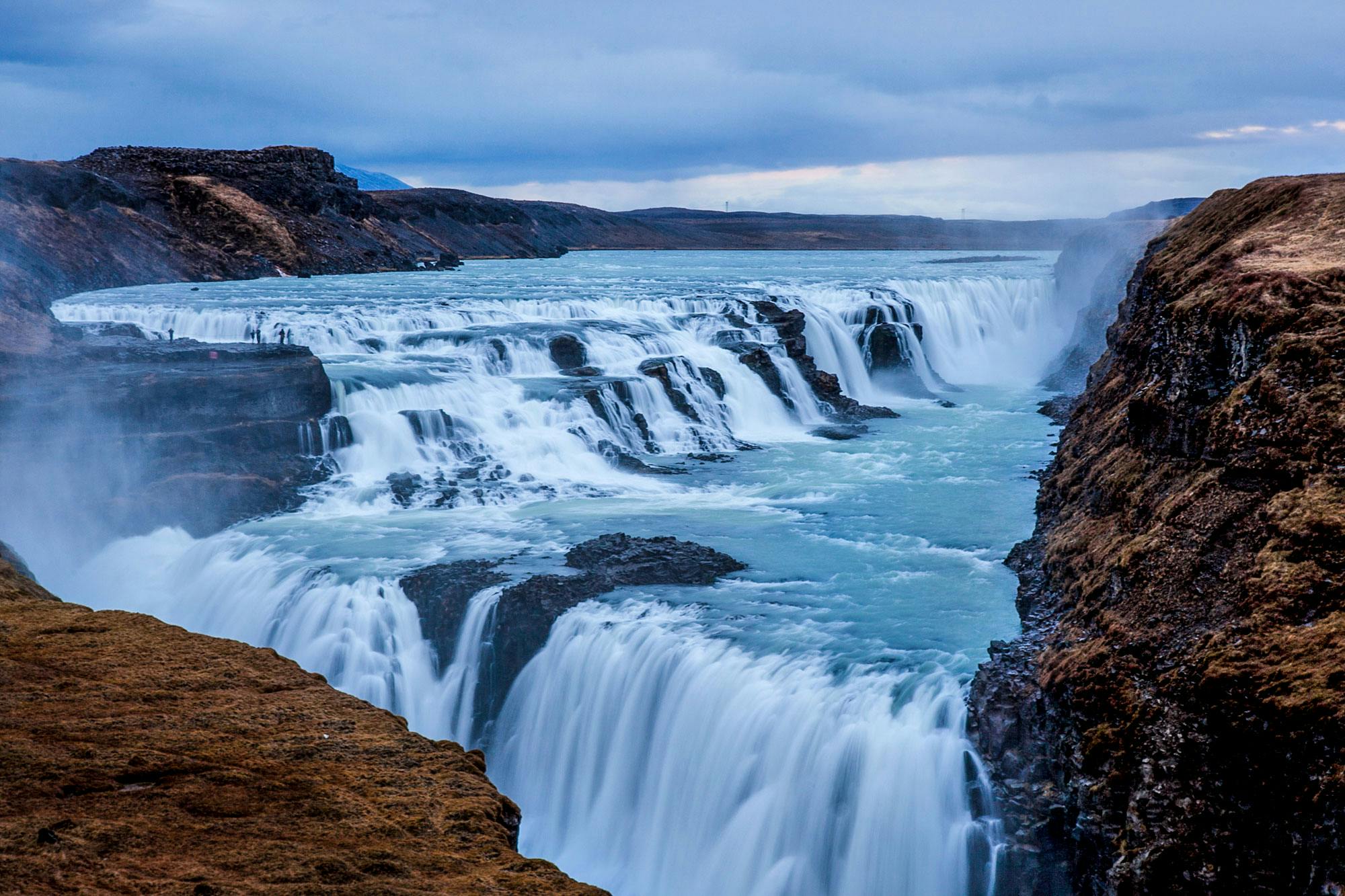 Wide, multi-tiered waterfall flowing through rocky terrain under a cloudy sky, with faint human figures visible on the left.
