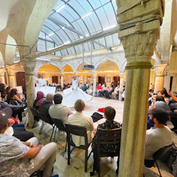 Människor som sitter i ett cirkulärt arrangemang runt virvlande dervischer som uppträder i en historisk hall med välvda pelare och takfönstertak.