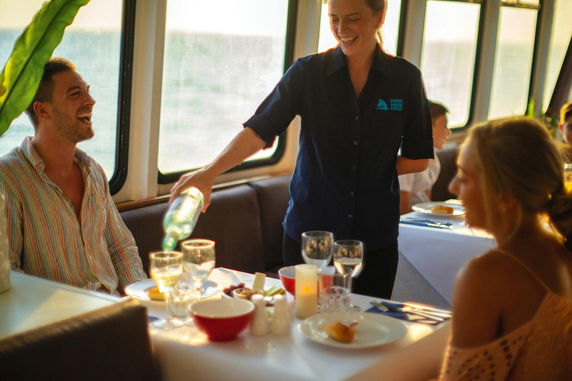 A smiling server in a blue shirt pouring wine for a seated diner in a well-lit dining area with tables set for a meal.