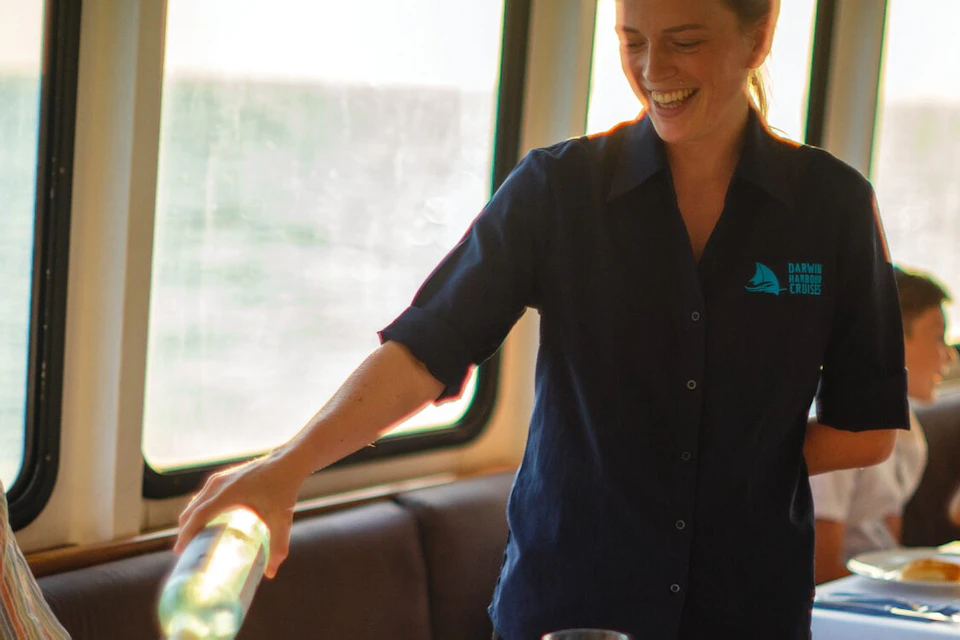 A smiling server in a blue shirt pouring wine for a seated diner in a well-lit dining area with tables set for a meal.
