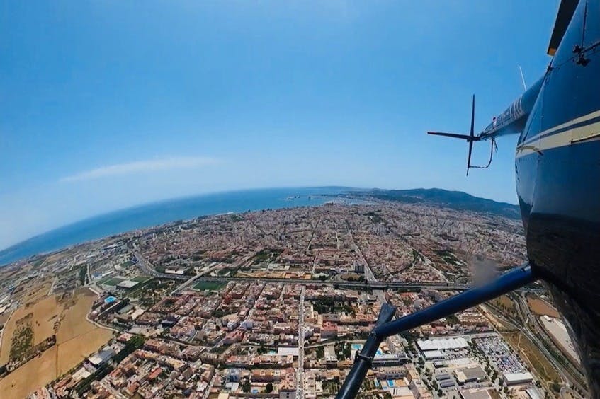 Αεροφωτογραφία παραλιακής πόλης με πυκνά κτίρια και ορατό στροφείο ελικοπτέρου σε πρώτο πλάνο, κάτω από καθαρό γαλάζιο ουρανό.