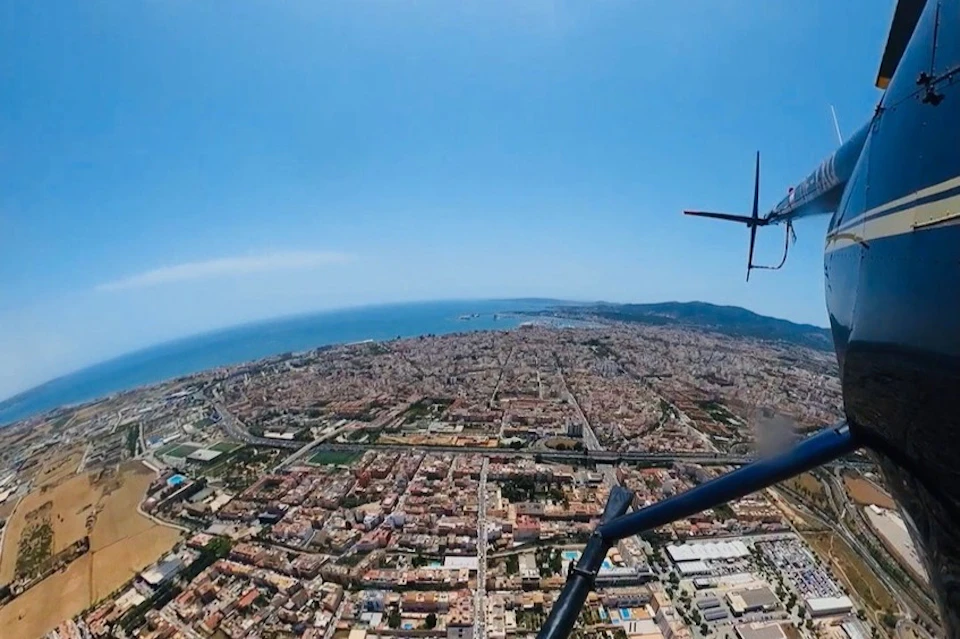 Aerial view of a coastal city with dense buildings and a visible helicopter rotor in the foreground, under a clear blue sky.