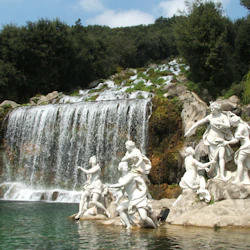White statues depicting human figures stand near a cascading waterfall surrounded by lush greenery.
