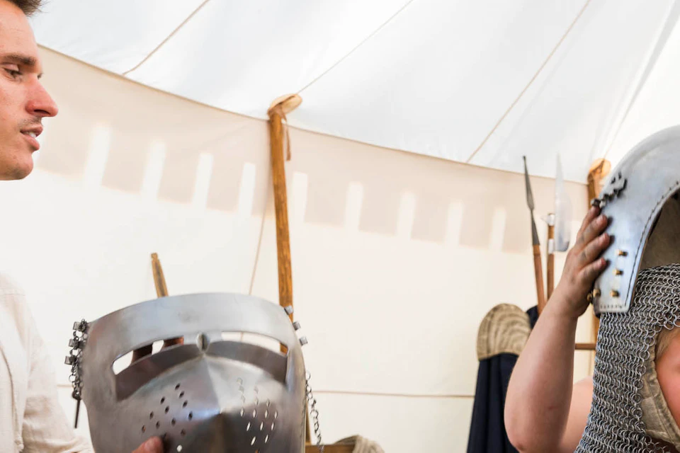 An adult helps a child put on a medieval knight's helmet inside a tent, with armor and medieval items displayed around.