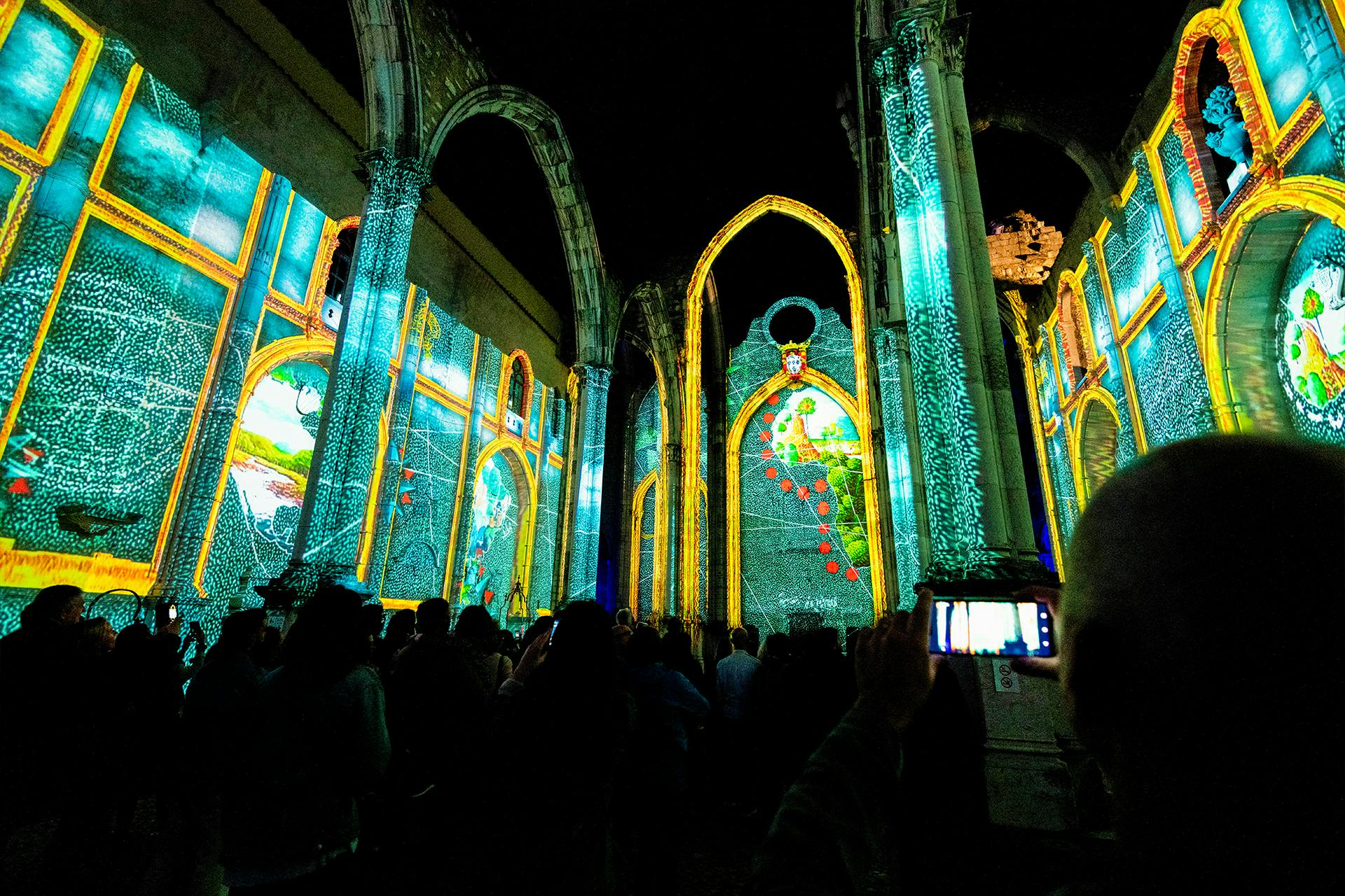 Crowd watches colorful projections on the interior walls of a historic, arched structure at night. A person records with a phone.