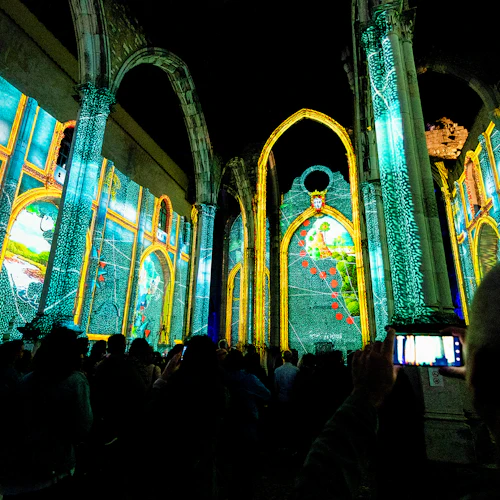 Crowd tittar på färgglada projektioner på innerväggarna i en historisk, välvd struktur på natten. En person spelar in med en telefon.