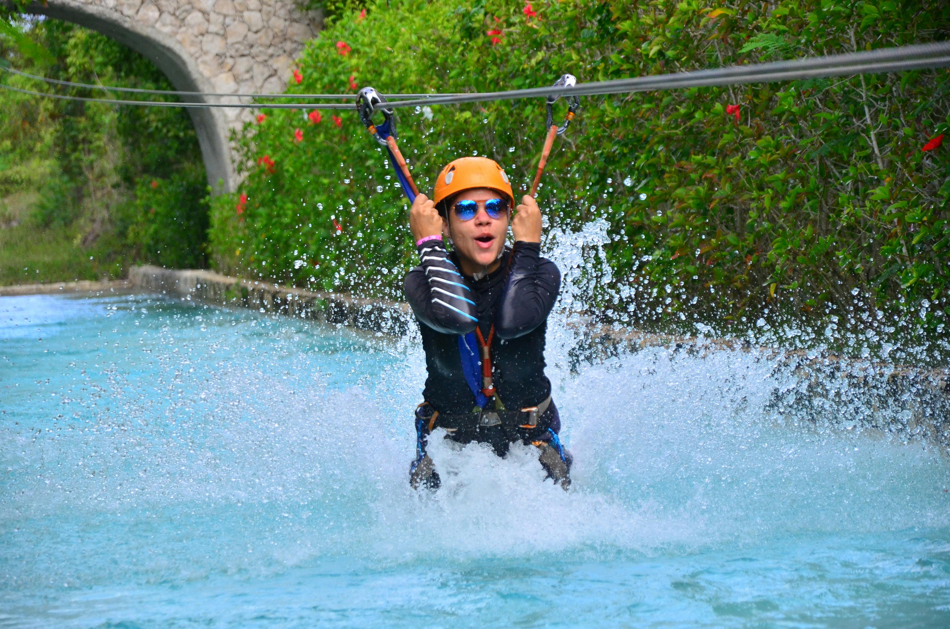 高空滑索上的人溅入游泳池，戴着头盔和太阳镜，背景是绿色植物和石拱门。