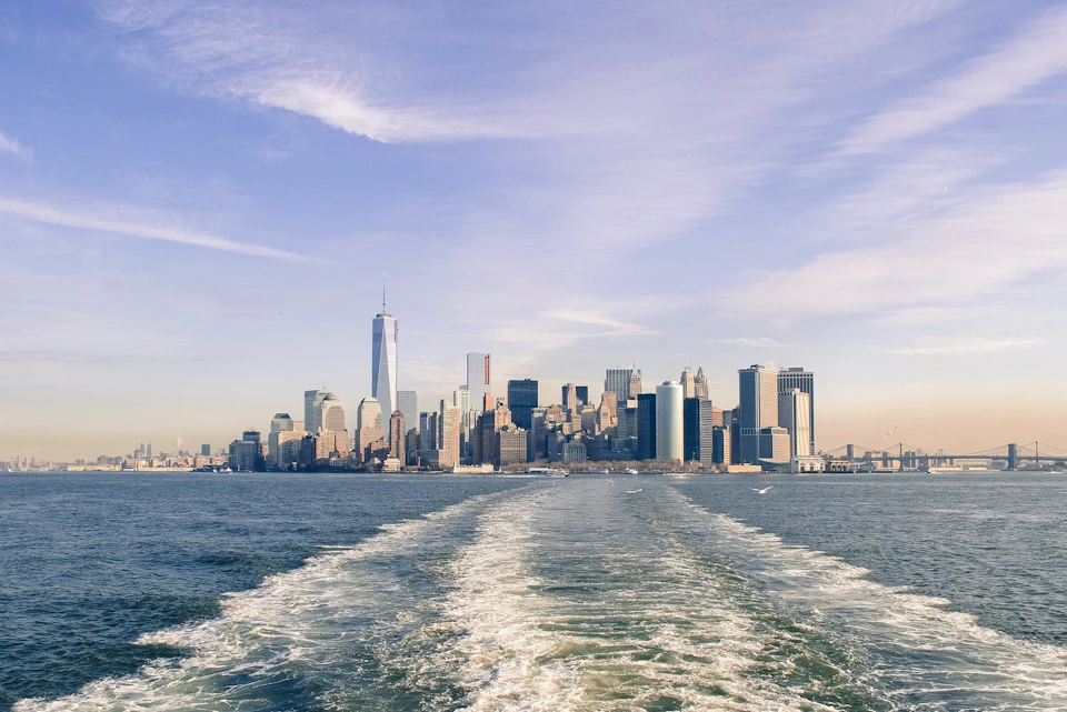 New York City skyline viewed from the water, with boat wake in the foreground and One World Trade Center prominently visible.