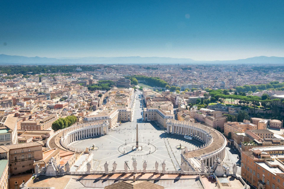 Aerial view of St. Peter's Square in Vatican City, featuring its colonnades, central obelisk, and surrounding urban landscape.