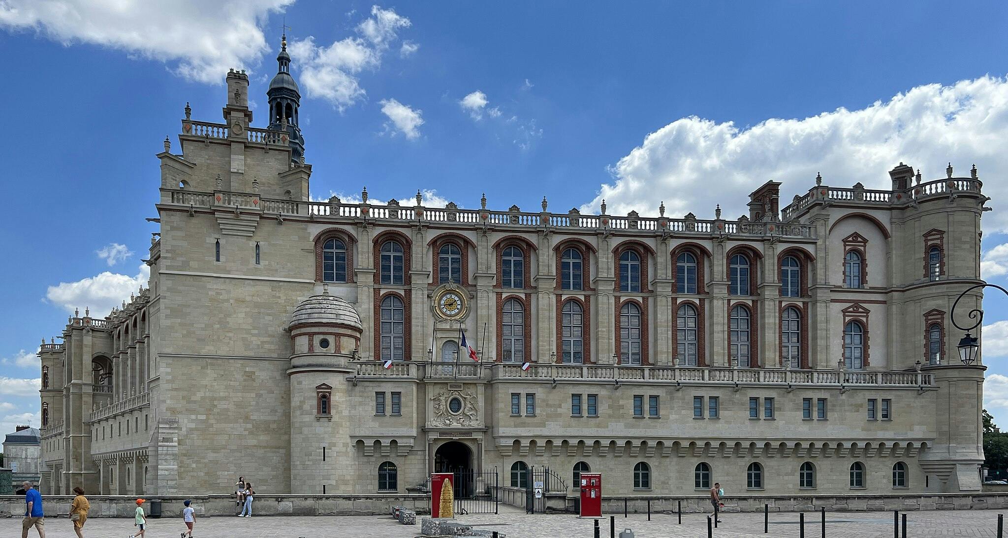 A historic stone building with a clock and flags above the entrance, set against a blue sky with a few clouds.