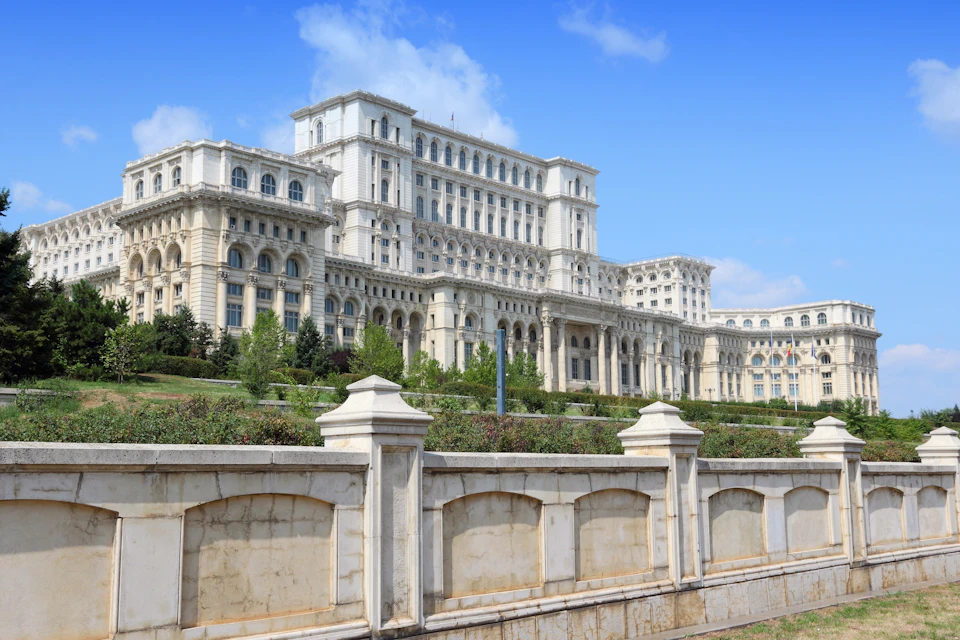 Large white neoclassical building with ornate architecture, surrounded by greenery and a stone fence, under a blue sky.
