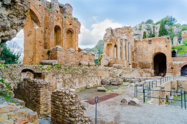 Ancient brick ruins with columns and arches, surrounded by rocky terrain and greenery under a partly cloudy sky.