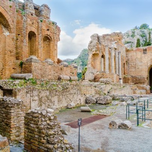 Ancient brick ruins with columns and arches, surrounded by rocky terrain and greenery under a partly cloudy sky.