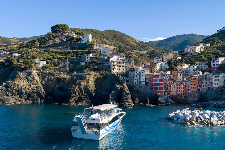 A coastal village with colorful buildings on rocky terrain, a tour boat in the foreground, and hills in the background.