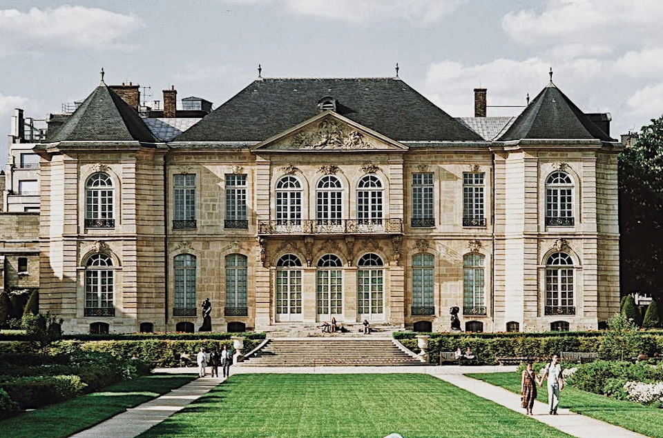 A grand historical building with symmetrical windows and a central balcony, set behind a manicured lawn with people walking.