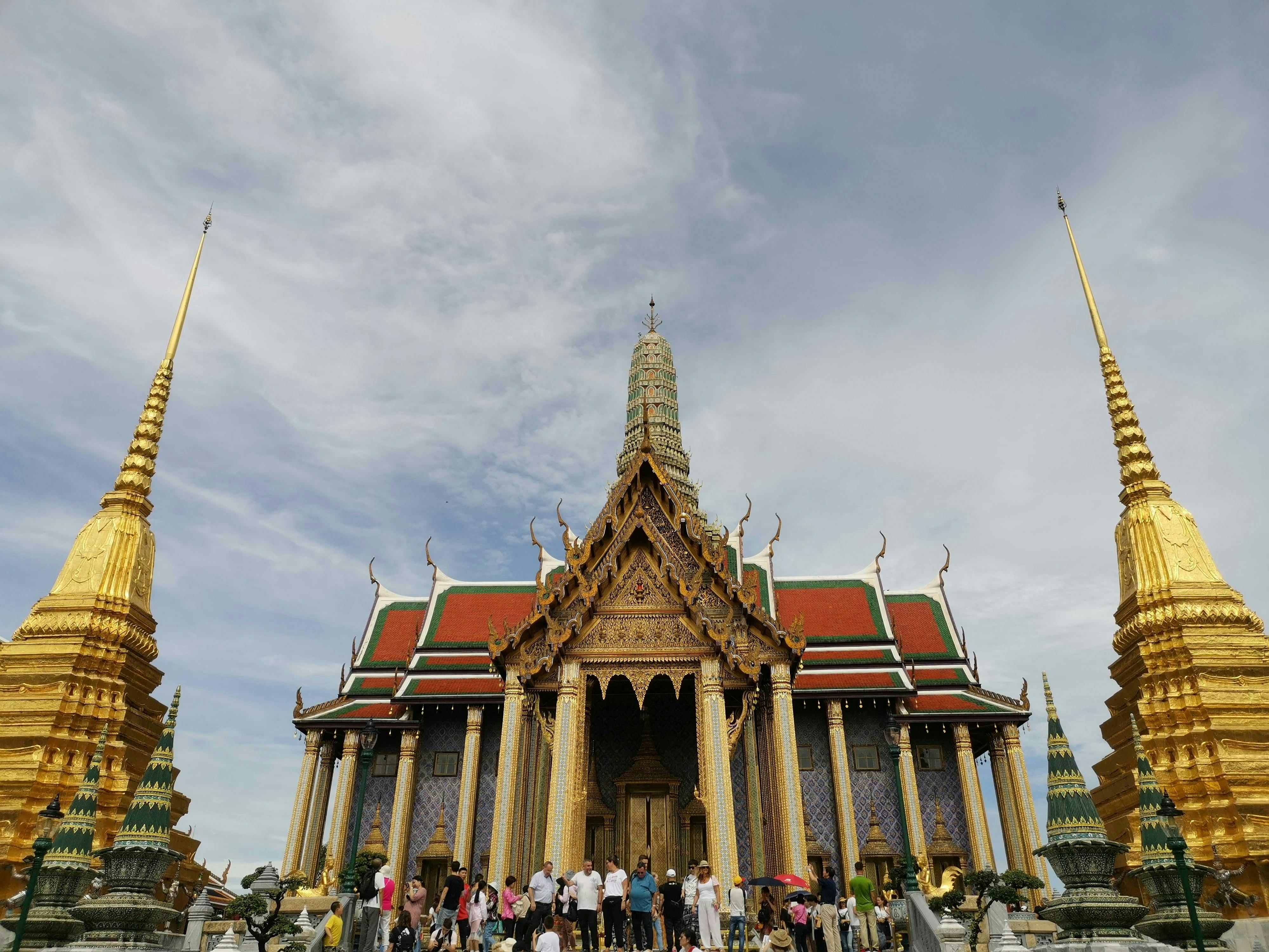 Os turistas visitam um templo ornamentado com pináculos dourados, pormenores intrincados e telhados coloridos sob um céu parcialmente nublado.