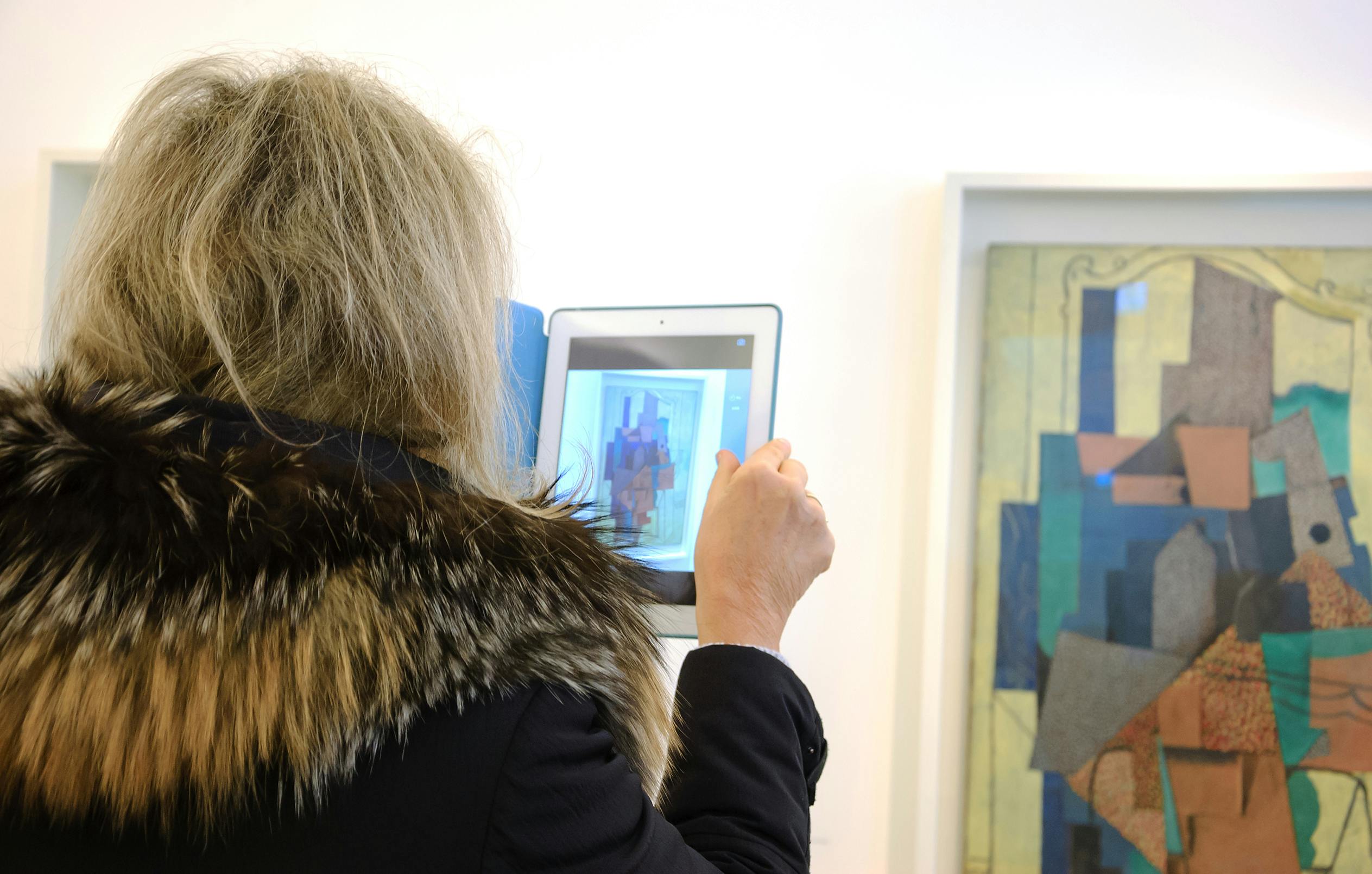 Person with long hair in a fur-trimmed coat photographing abstract art with a tablet in a gallery.