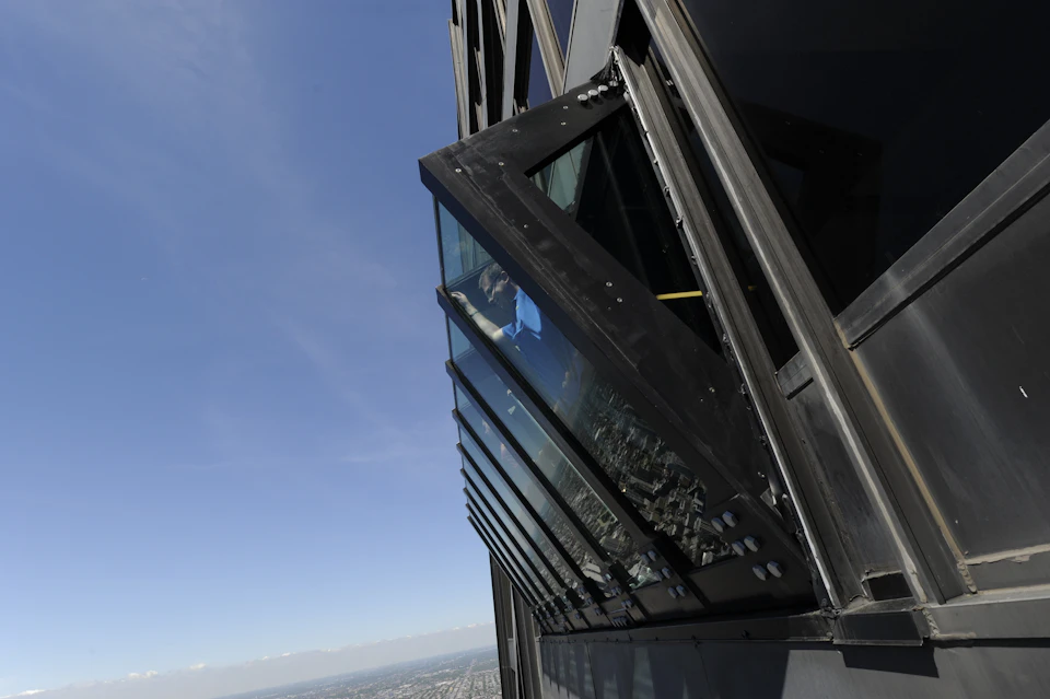 A person stands inside a tilted glass window box high above a cityscape, with a clear blue sky in the background.