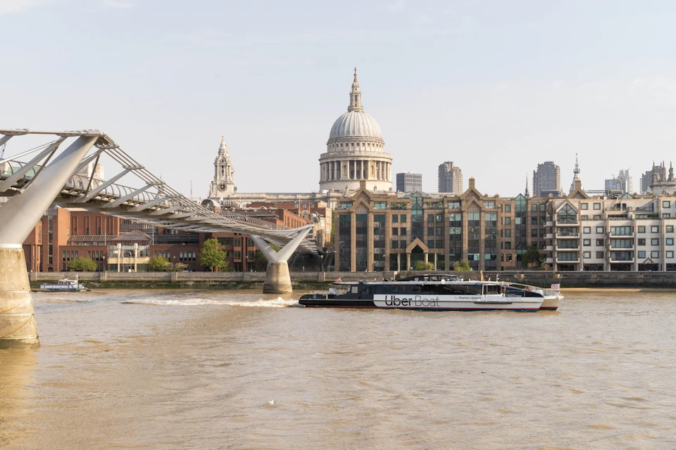 A view of St. Paul's Cathedral, the Millennium Bridge, and an Uber Boat on the Thames in London under a clear sky.