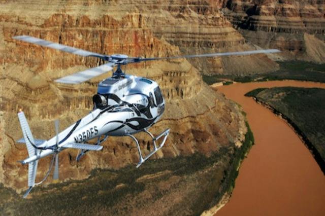 A helicopter flies over a canyon with steep, layered rock formations and a winding muddy river below.