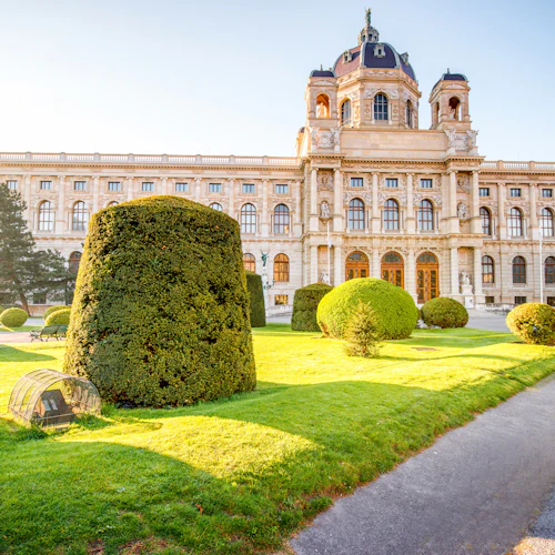 En stor, historisk byggnad med välvda fönster, en kupol och prydligt trimmade häckar i ett solbelyst trädgårdsområde.