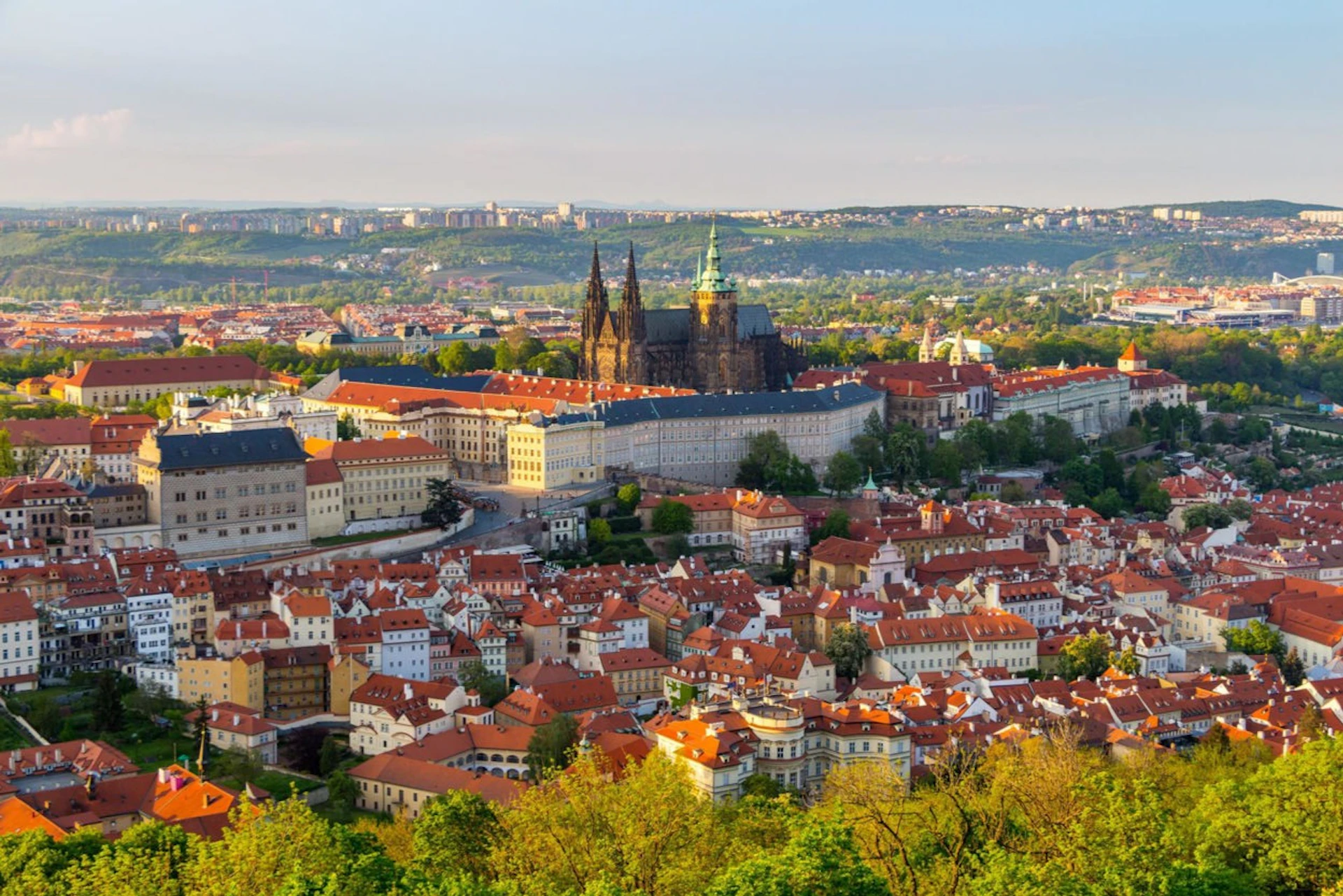 A panoramic view of a city with red-roofed buildings, a cathedral, and greenery under a clear sky.