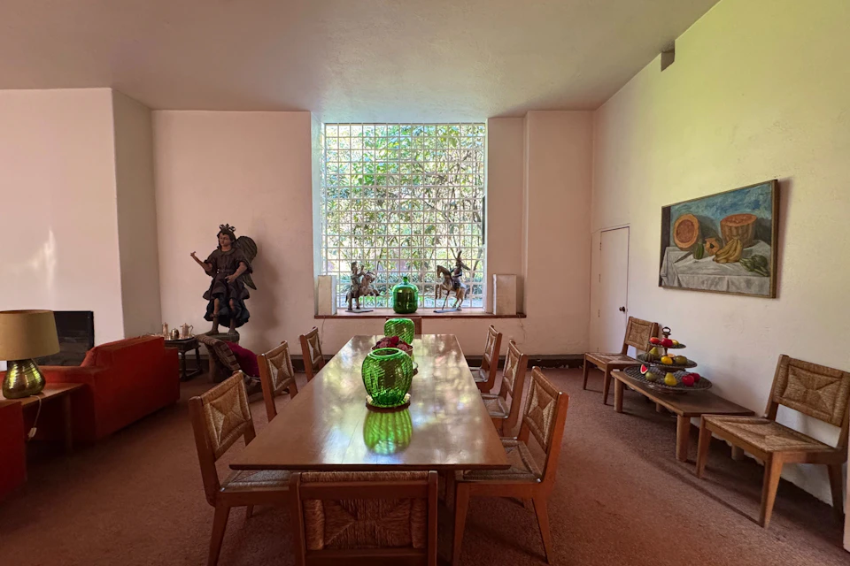 A room with a wooden table, green glass vases, wicker chairs, sculptures, a window with grid glass, and a colorful wall painting.
