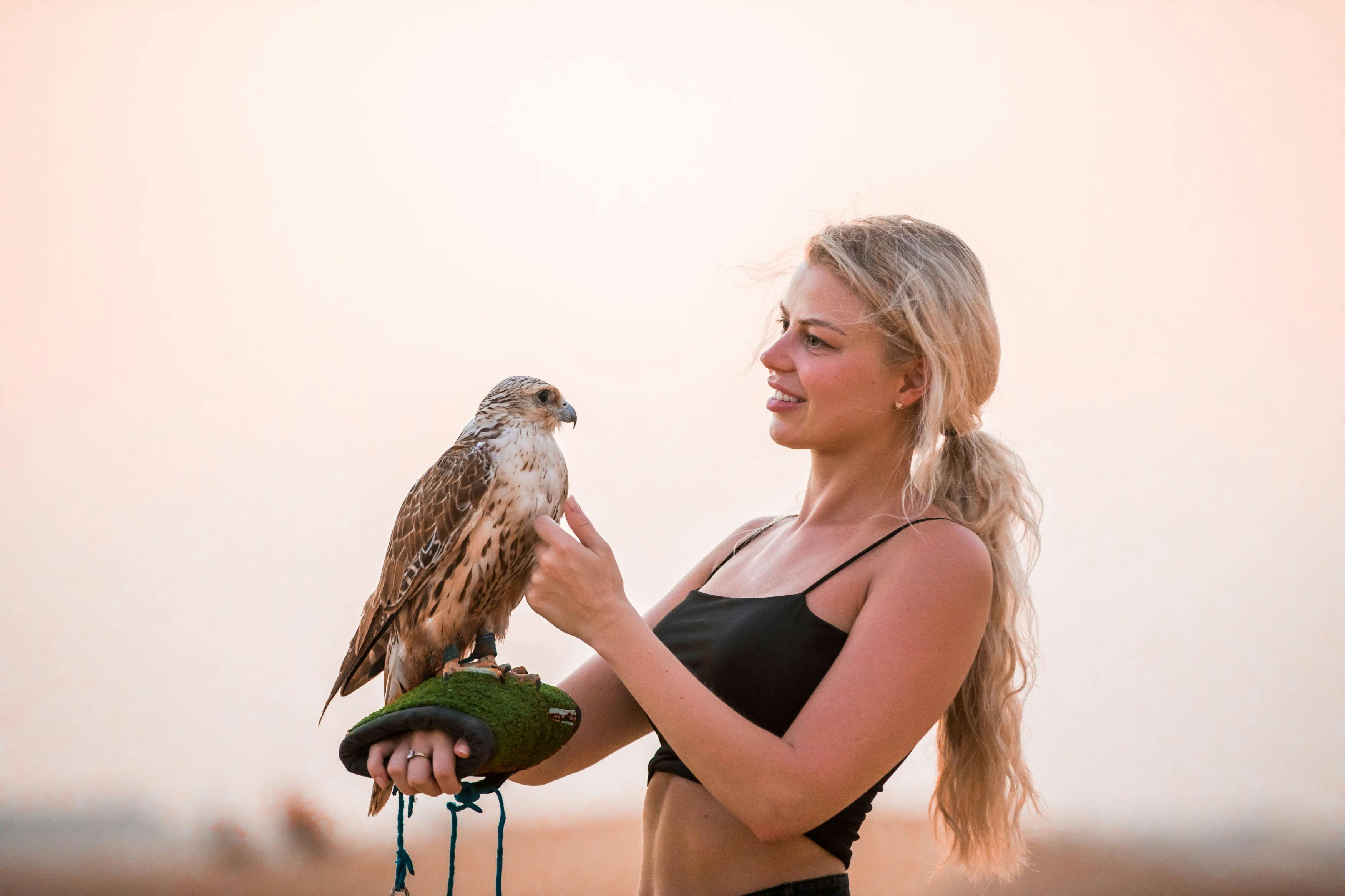 A woman with long blonde hair holds a falcon on a gloved hand against a soft, peach-colored sky background.