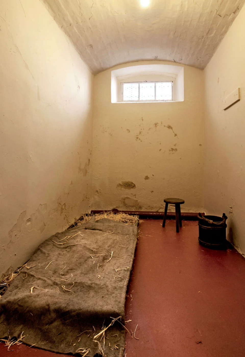 Small, bare room with peeling paint, a small bed with straw, a stool, a bucket, and a small window near the ceiling.