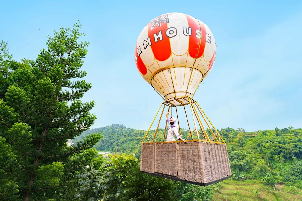 A hot air balloon with a red and white striped envelope labeled "FARMHOUSE" carrying a wicker basket with a person inside.