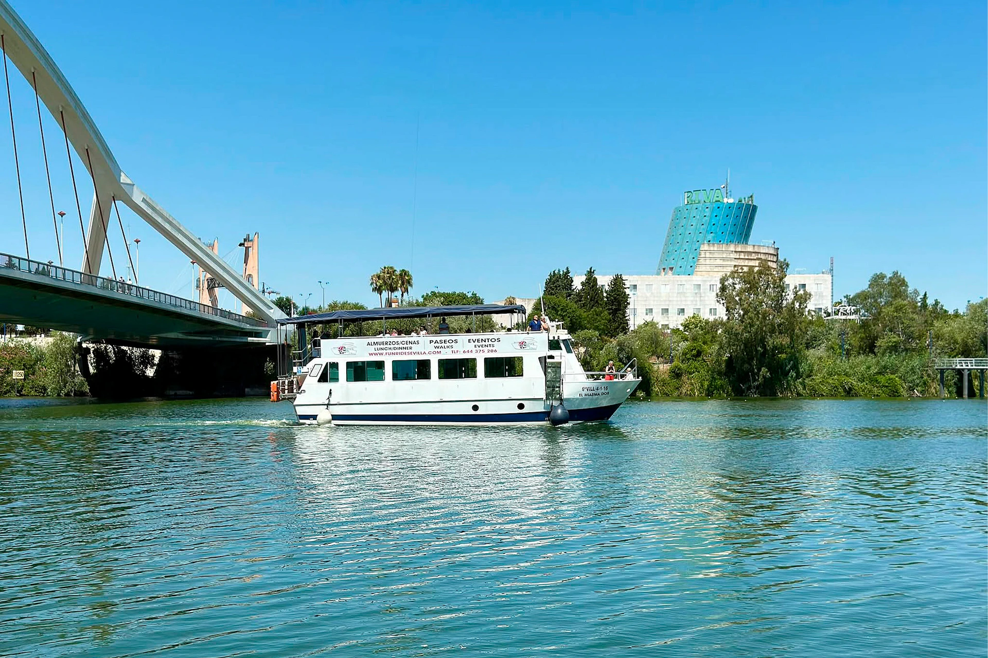 A white passenger boat on a river, with a bridge, trees and buildings in the background, beneath a clear blue sky.