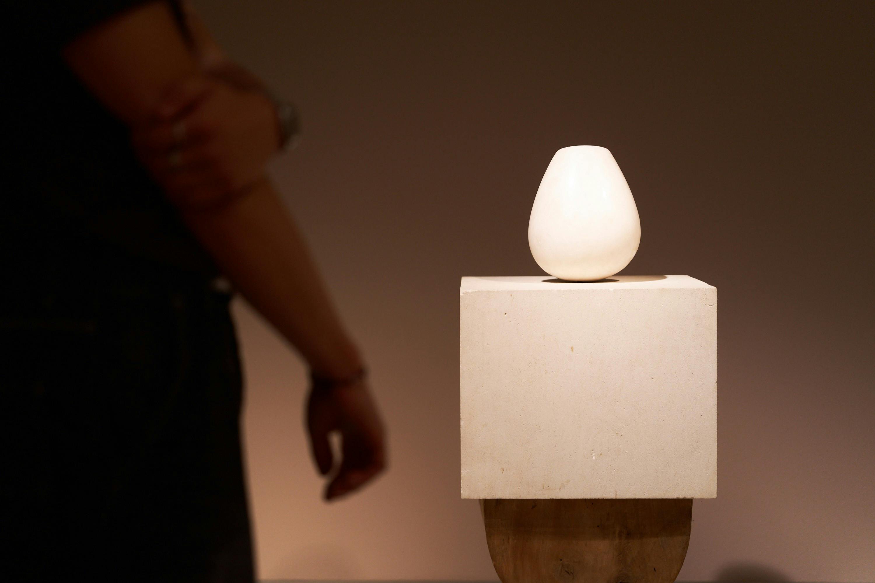 Ένα λευκό γλυπτό σε σχήμα αυγού σε ένα βάθρο με ένα άτομο να στέκεται στο παρασκήνιο.