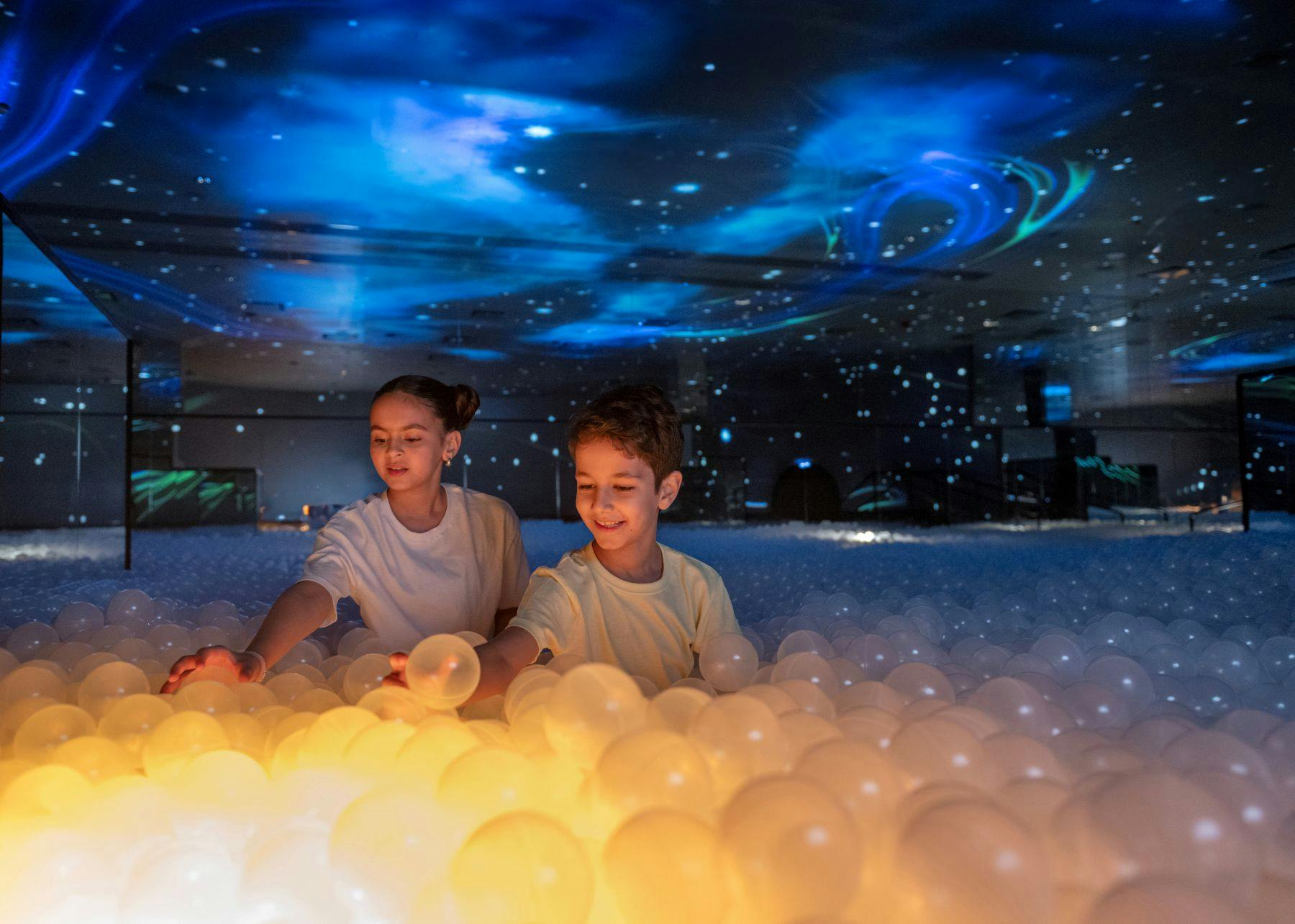 어둡고 별이 빛나는 분위기 있는 천장 아래 빛나는 공이 가득한 수영장에서 놀고 있는 두 어린이.