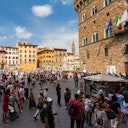 A bustling square with tourists, historic buildings, souvenir stalls, and flags on a stone building under a blue sky.