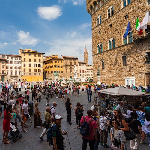 A bustling square with tourists, historic buildings, souvenir stalls, and flags on a stone building under a blue sky.